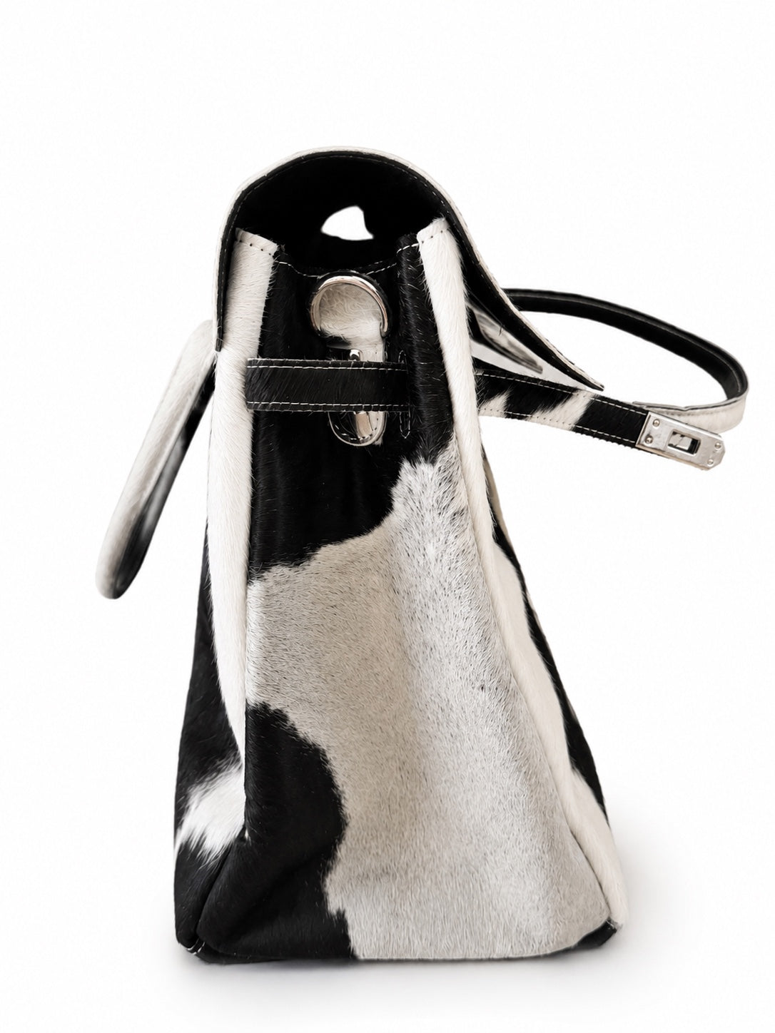 A structured handbag with a black and white cowhide pattern, featuring a black leather strap with silver-tone hardware and a silver circular accent on the front.