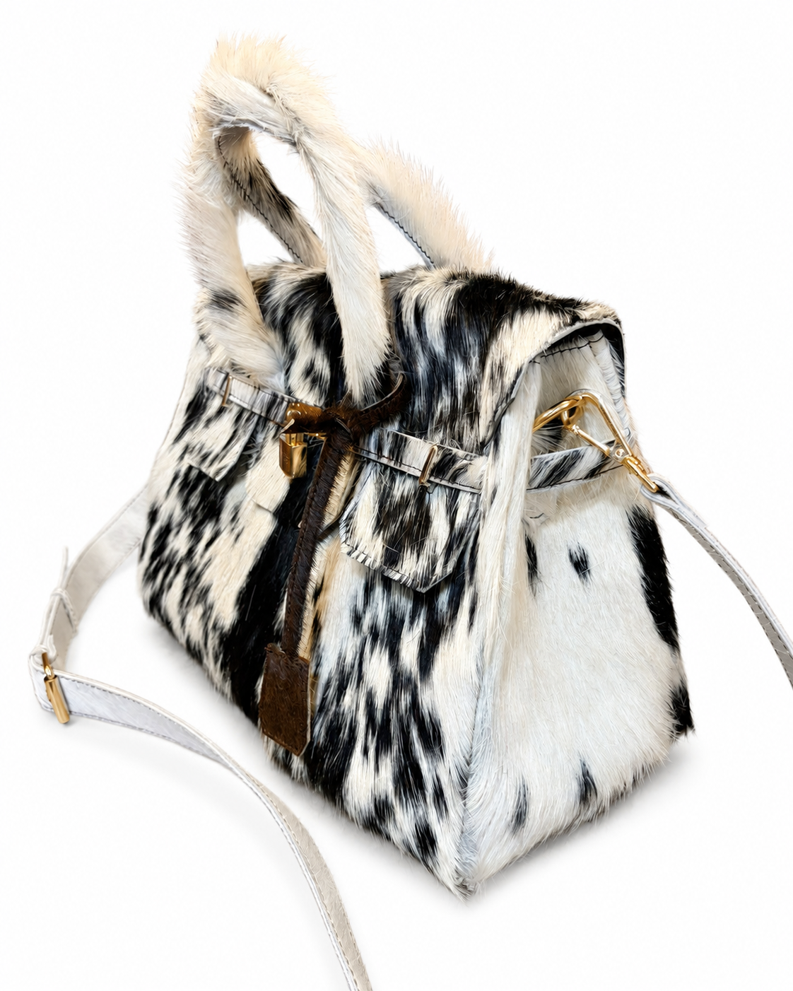 A structured handbag with a black and white cowhide pattern, featuring faux fur trim on the handles and a brown leather strap detail with gold-tone hardware, shown on a plain white background.