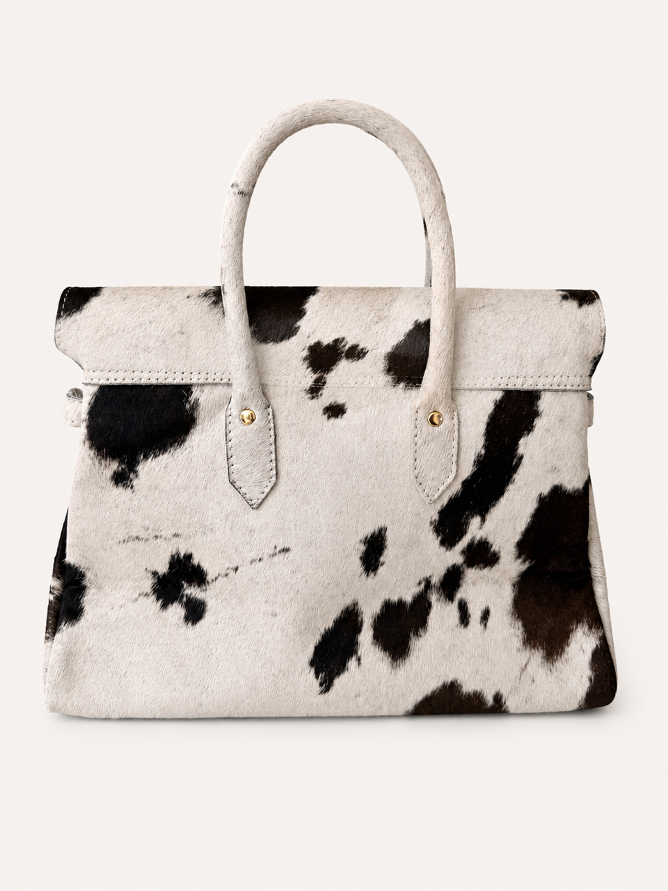 A structured handbag with a black and white cowhide pattern, featuring dual top handles and gold-tone hardware, set against a plain off-white background.