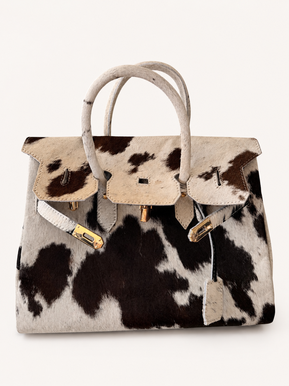 A structured handbag with a cowhide pattern featuring black and brown spots on a cream background, with beige leather trim, double top handles, and gold-tone hardware, shown against a plain off-white background.