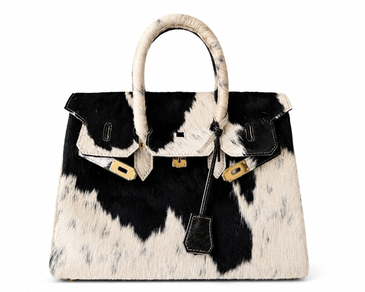 A structured handbag with a black and white cowhide pattern, featuring a single rolled top handle, gold-tone hardware, and a hanging leather clochette, photographed against a plain white background.
