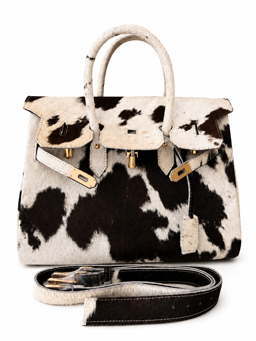 A structured handbag with a black and white cowhide pattern, featuring cream-colored leather handles and trim, gold-tone hardware, and a matching detachable cowhide shoulder strap placed in front on a white background.