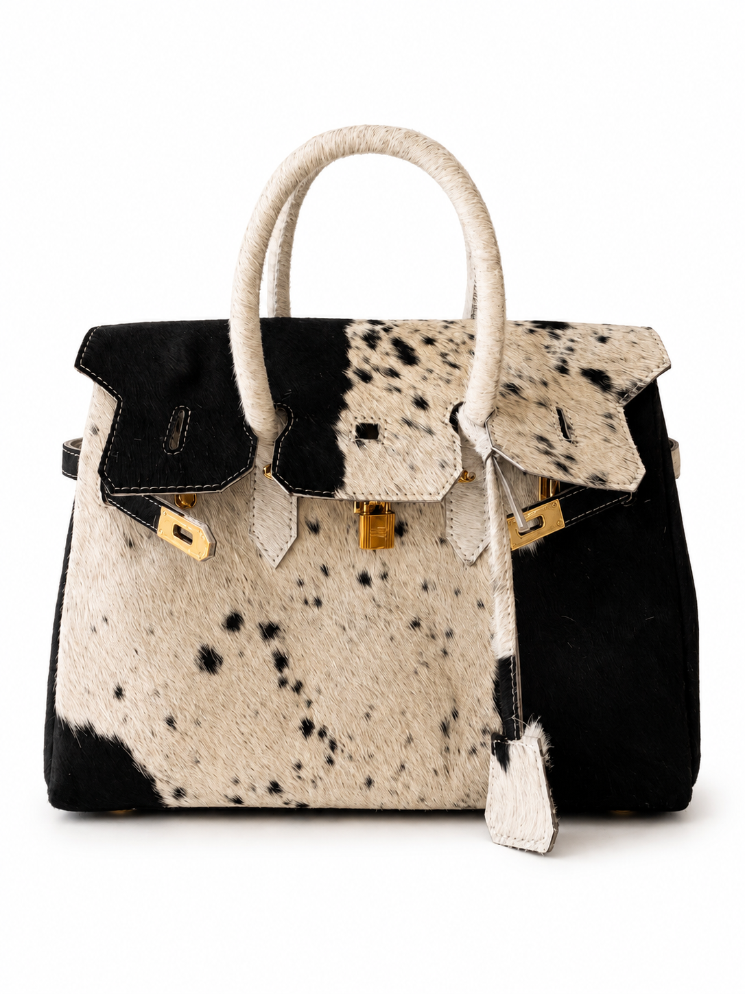 A structured handbag with a split black and black-and-white cowhide pattern, featuring cream-colored handles, gold-tone hardware, and a hanging leather clochette, photographed on a clean white background.
